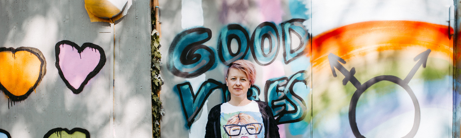 Sabrina Paulino steht vor einer Graffitiwand mit dem Schriftzug good vibes und dem Symbol für geschlechtliche Vielfalt.