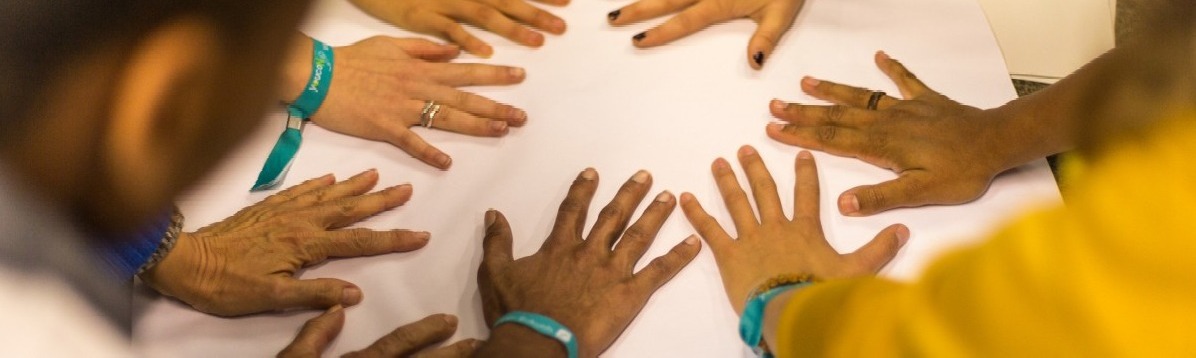 Mehrere junge Menschen strecken ihre Hände im Kreis auf ein weißes Blatt. Sie tragen türkise youcoN-Festivalbändchen